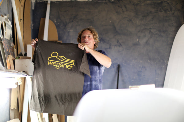 John Wegener standing in his surfboard shaping room holding his Wegener Surfboards brown surf t-shirt