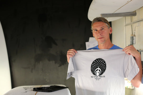 Michael Miller surfboard shaper standing in his shaping room holding his Peacock surf t-shirt